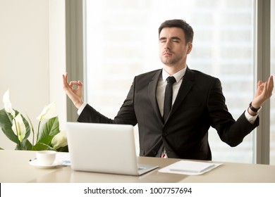 Handsome Man In Business Suit Meditating With Closed Eyes At Work Desk With Laptop. Successful Businessman Practicing Eastern Spiritual Practices At Office Workplace For Relaxing And Stress Relief