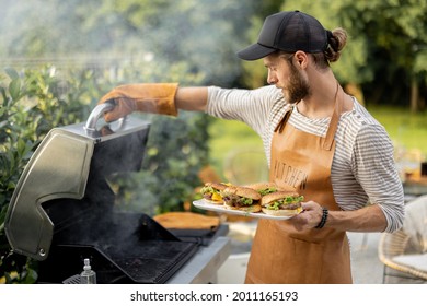 Handsome Man In Cap And Apron Making Burgers On A Grill At Backyard. Cooking Outdoors And American Lifestyle Concept