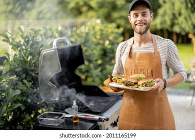 Handsome man in cap and apron making burgers on a grill at backyard. Cooking outdoors and american lifestyle concept - Powered by Shutterstock