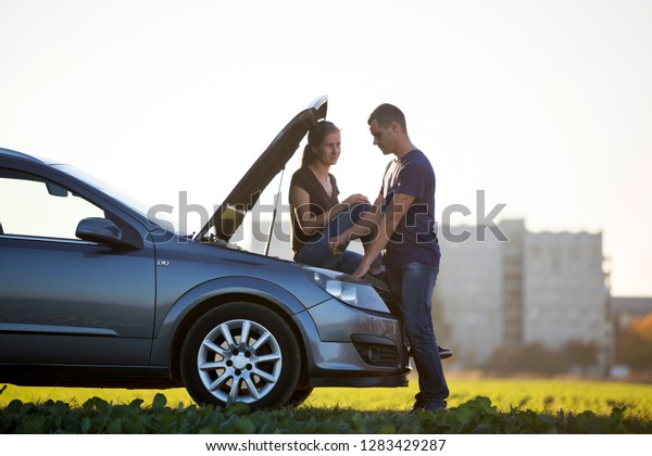 Handsome man at car with popped hood checking\
oil level in engine using dipstick and attractive woman watching on\
clear sky background. Transportation, vehicles problems and\
breakdowns concept.