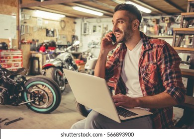 Handsome man in casual clothes is talking on the mobile phone, using a laptop and smiling while sitting in the motorcycle repair shop - Powered by Shutterstock