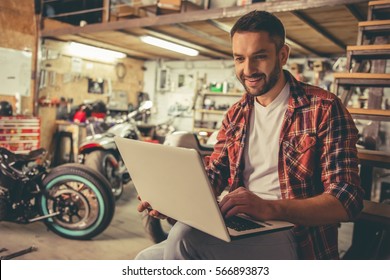 Handsome man in casual clothes is using a laptop and smiling while sitting in the motorcycle repair shop - Powered by Shutterstock