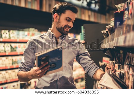 Handsome man is choosing a magazine and smiling while doing shopping at the supermarket