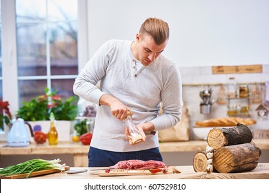 Handsome Man Cooking At Home Preparing Meat Stake From Pork Or Beef Or Lamb, In Authentic Modern Light Kitchen With Wooden Surface, Full Of Fancy Kitchenware