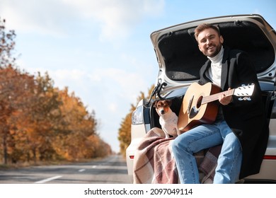 Handsome Man With Cute Dog Playing Guitar In Car Trunk On Autumn Day