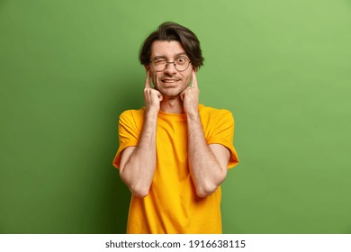 Handsome Man With Dark Hair Plugs Ears Winks Eye Avoids Loud Sound Wears Casual Yellow T Shirt Round Spectacles Isolated Over Green Background. Its Too Noisy Here. Hipster Guy Refusing To Listen