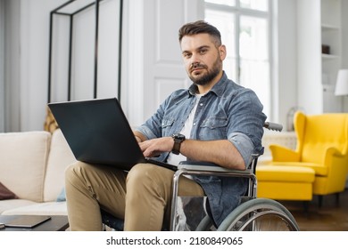 Handsome Man With A Disability Sitting In Wheelchair With Laptop On Knees And Looking At Camera. Caucasian Male In Casual Wear Having Opportunities To Remote Work At Home.
