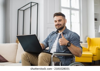 Handsome Man With A Disability Sitting In Wheelchair With Laptop On Knees And Looking At Camera. Caucasian Male In Casual Wear Having Opportunities To Remote Work At Home Showing Thumb Up.