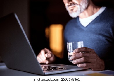 Handsome man drinking while working from home office at evening, using laptop. Screen glow illuminates man's face. Telecommuting and home office.