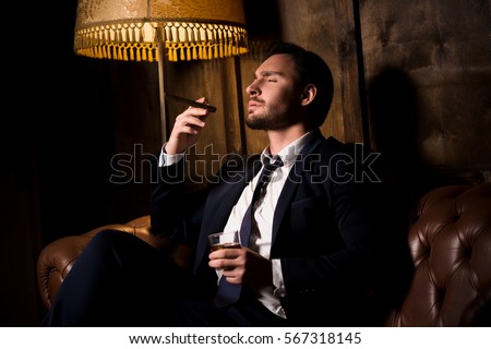 Handsome man drinking whiskey and smoking cigar. Billionaire man resting and relaxing while sitting on sofa in men's club. Luxury concept.