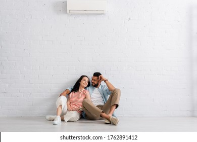 Handsome man embracing beautiful girlfriend while sitting under air conditioner on floor at home