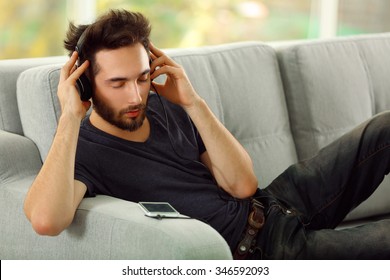 Handsome man emotionally listens music with headphones on grey sofa in the room