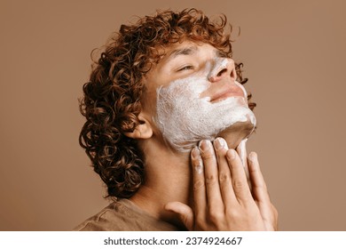 Handsome man enjoying beauty treatment while applying face cream and standing on brown background - Powered by Shutterstock