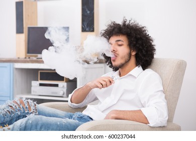 Handsome Man Enjoying Free Time On Weekends. Hipster Man Sitting In Arm Chair And Smoking While Staying At Home.