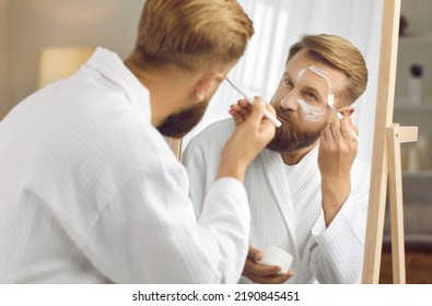 Handsome Man Enjoying Spa Day At Home. Attractive Adult Metrosexual In Clean White Bathrobe Looking In Bathroom Mirror And Applying White Kaolin Clay Mask On Face. Male Beauty And Skin Care Concept