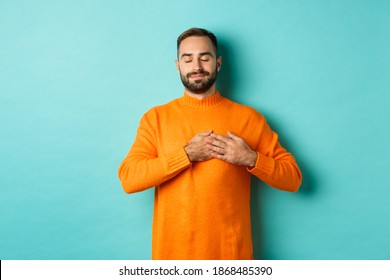 Handsome man feeling nostalgic, holding hands on heart and daydreaming, remember or imaging something, standing carefree against turquoise background