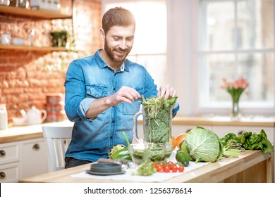 Handsome man filling blender with green raw food, making vegetarian smoothie on the kitchen at home - Powered by Shutterstock