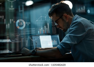 Handsome Man In Glasses And Shirt Writing In Notepad Sitting At Desk With Laptop In Empty Dark Office. 