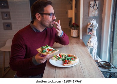 Handsome Man With Glasses Sitting At Restaurant Holding Slice Of Pizza And Licking His Fingers.