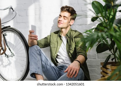 Handsome Man In Green Shirt Holding Smartphone, Sitting Near Brick Wall, Bicycle And Plants In Office