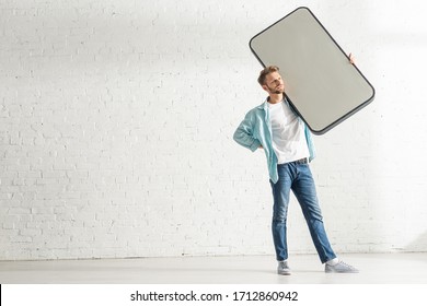 Handsome man with hand on hip holding big model of smartphone near white brick wall - Powered by Shutterstock