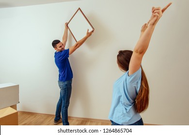 Handsome Man Hanging Painting In Living Room While His Girlfriend Having Fun. Young Family Moving In.