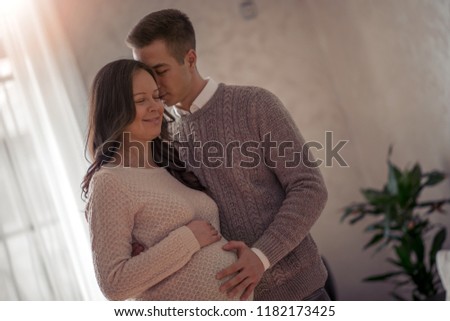 Handsome man and his beautiful pregnant wife are hugging and smiling while standing near the window at home.