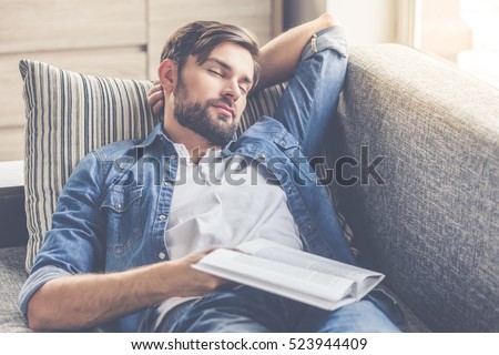 Handsome man is holding a book and napping while lying on couch at home