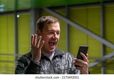 Handsome man holding mobile phone, screaming, celebrating good news while standing in covered passage. Enthusiastic young man holding mobile phone and celebrating win outdoors.  - Shutterstock ID 2173492051