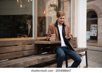 Handsome Man Holding Takeaway Coffee Cup And Mobile Phone In Hand. City Life Cafe In Background. Man In Brown Coat Modern Businessman With Backpack