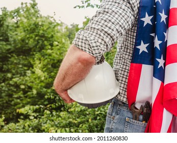Handsome man holding US Flag and construction helmet against the background of trees, blue sky and sunset. View from the back. Labor and employment concept - Shutterstock ID 2011260830