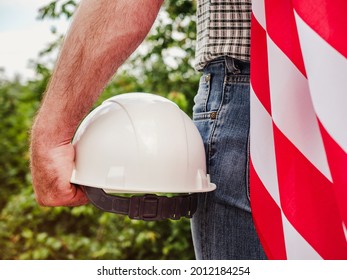 Handsome man holding US Flag and construction helmet against the background of trees, blue sky and sunset. View from the back. Labor and employment concept - Shutterstock ID 2012184254