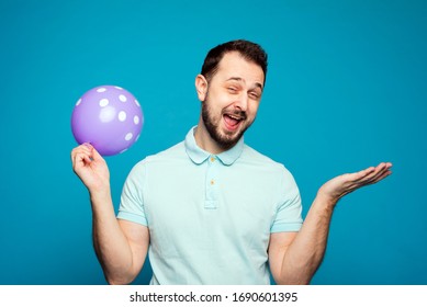 A Handsome Man Holds A Balloon In His Hand And Rejoices And Screams At A Holiday Or Birthday. A Man Inhaled Laughing Gas, Helium And Laughs On A Blue Background In The Studio