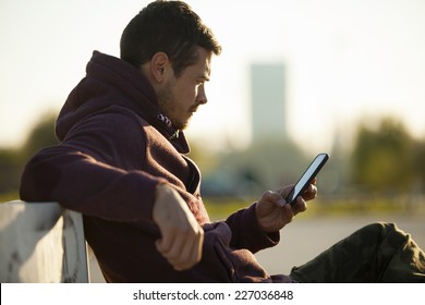 Handsome Man In Hoodie Holding And Looking At Mobile Smart Phone Outdoors