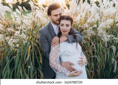 Handsome Man Hugging Beautiful Pregnant Woman In White Dress At Uruguayan Pampas Grass Background In Evening 