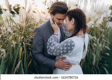 Handsome Man Hugging Beautiful Pregnant Woman In White Dress At Uruguayan Pampas Grass Background In Evening 