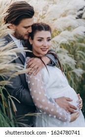 Handsome Man Hugging Beautiful Pregnant Woman In White Dress At Uruguayan Pampas Grass Background In Evening 