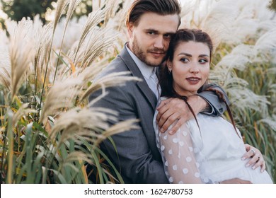 Handsome Man Hugging Beautiful Pregnant Woman In White Dress At Uruguayan Pampas Grass Background In Evening 