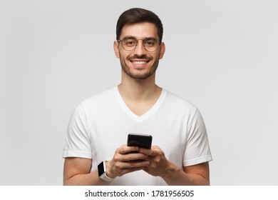 Handsome Man Isolated On Gray Background, Looking Straight At Camera While Holding His Phone