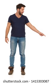 Handsome man in jeans, boots and blue t-shirt is standing, pointing down and showing something. Full length studio shot isolated on white.