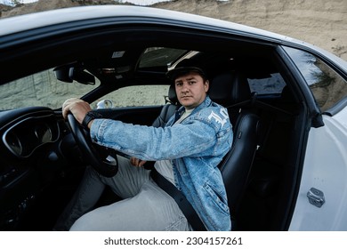 Handsome man in jeans jacket and cap sit at his white muscle car.