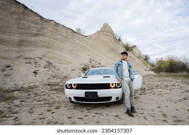 Handsome man in jeans jacket and cap is standing near his white muscle car in career.