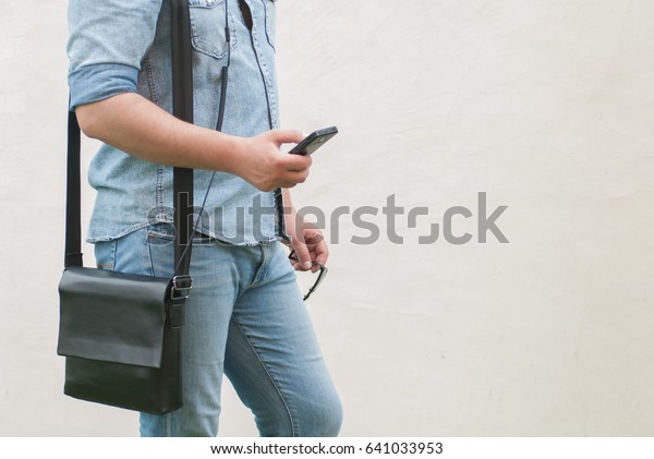 Handsome man in jeans jacket, hipster\
with shoulder bag stays from the left and holds a phone in hand,\
photo with no face, white copy space for text from the\
right