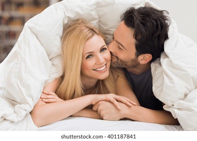 Handsome Man Kissing Woman On Cheek Under A Duvet In The Bed. Happy Young Couple Lying Down Comfortably In Their Bed At Home. Young Couple Having Fun Lying On Bed At Home.
