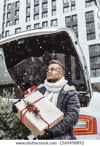 Handsome man in knitted scarf and black gloves holding a gift box, is waiting for his beloved. Urban background. Waiting near the car in snowfall.