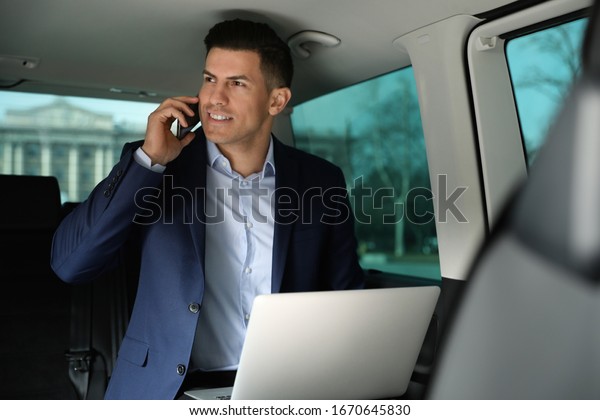 Handsome man with laptop talking on smartphone in\
modern car