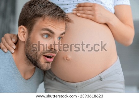 Handsome man is listening to his beautiful pregnant wife's tummy and excited. Future father feeling hearing first bumps of baby embryo fetus. Parenting