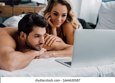 Handsome man looking at laptop near smiling woman on bed
