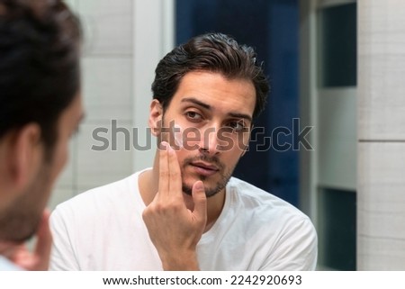 Handsome man looking at mirror and applying moisturizing cream on cheeks in bathroom. Groomed young guy doing skincare morning routine after taking a shower