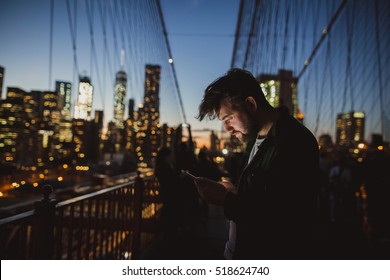 Handsome Man Looking At Mobile Phone At Brooklyn Bridge At Night. Amazing View Of New York On The Background.